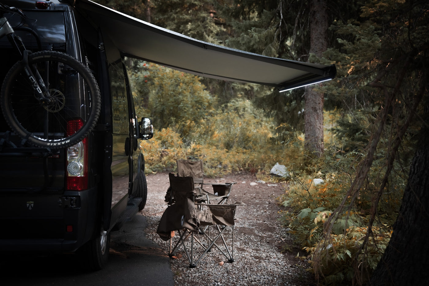 Relaxing rv rental campsite scene: dark camper van with bike, extended awning, and camp chairs among trees. — photo 5