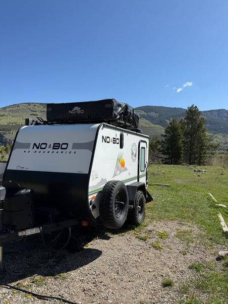 A white NO BO adventure rv rental trailer with a rooftop tent, parked on gravel in a mountain landscape under a clear blue sky.