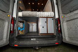 Inside an RV rental camper van: rear view showing a bed, built-in storage, and the electrical system on the left.