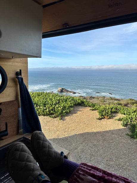 Relaxing in an RV rental, feet up, enjoying the incredible ocean view with sea stacks & lush coastal plants from inside the van.