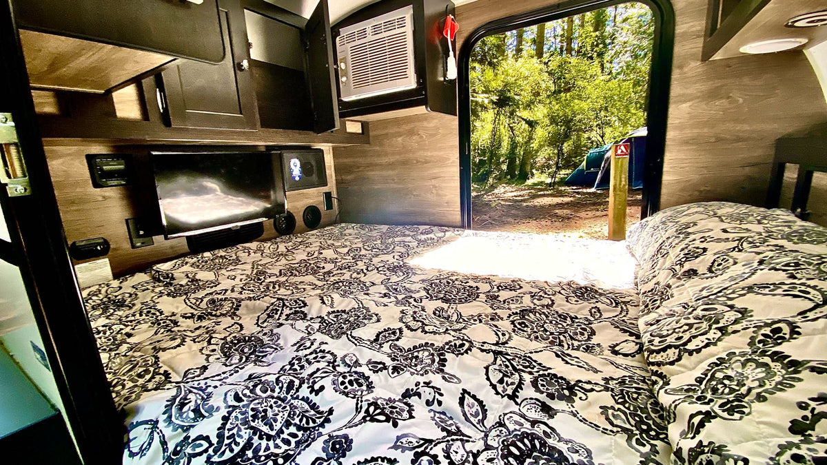 Cozy RV rental interior: bed, TV, & AC. The open door shows a forest campsite with a blue tent & wooden post. — photo 5