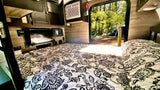 Cozy RV rental interior: bed, TV, & AC. The open door shows a forest campsite with a blue tent & wooden post.
