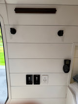 RV rental interior: White shiplap wall features modern light controls, USB outlet, MAXAIR remote, hooks, and a dark shelf.