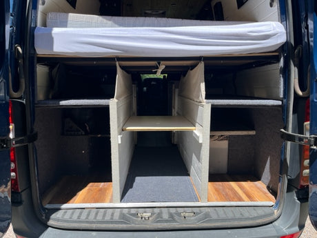 Rear view of an RV rental camper van showing an organized interior with an elevated bed, ample storage bays, and a central aisle.