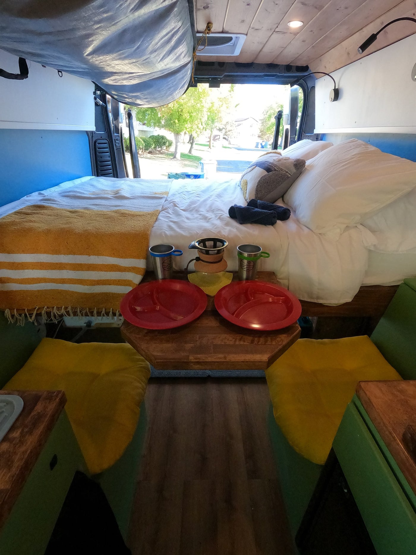 Inside this RV rental, a comfy bed and dining setup with plates and coffee maker. Open rear doors reveal an outdoor view. — photo 12