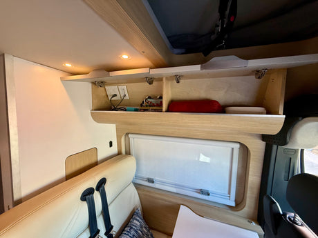 Open overhead storage with power outlets, a window, and comfortable beige seating with seatbelts in an RV rental.