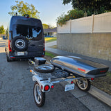 Dark gray Mercedes Sprinter RV rental van towing a compact trailer with a Yakima SkyBox cargo box and a spare tire.