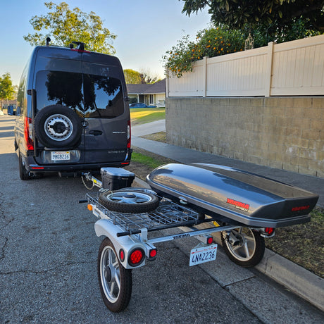 Dark gray Mercedes Sprinter RV rental van towing a compact trailer with a Yakima SkyBox cargo box and a spare tire.