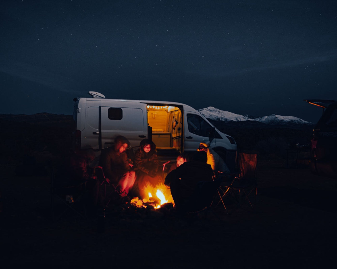 RV rental fun: Friends gathered by a glowing campfire, under a starry sky, with a camper van & snowy peaks. — photo 4