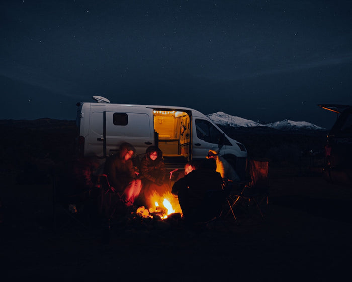 RV rental fun: Friends gathered by a glowing campfire, under a starry sky, with a camper van & snowy peaks.
