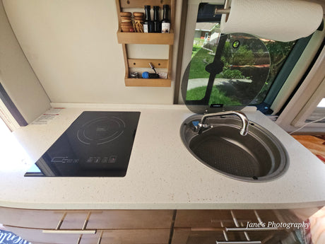 RV rental kitchen: light-speckled counter with black induction cooktop, covered oval sink, and wooden wall shelves for storage.