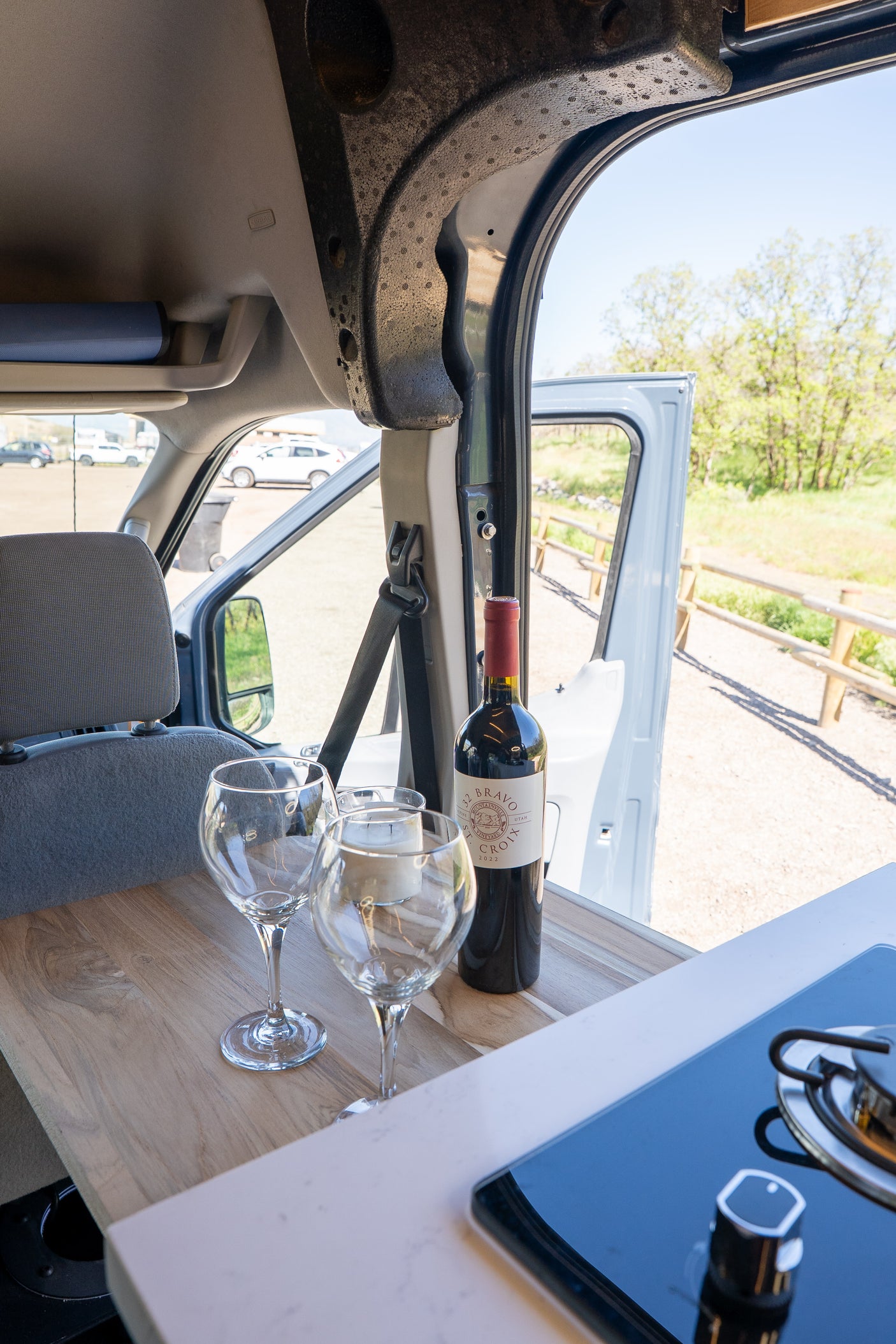 RV rental interior: Wine, glasses, and a stovetop inside, with an open door revealing trees & a wooden fence outside.