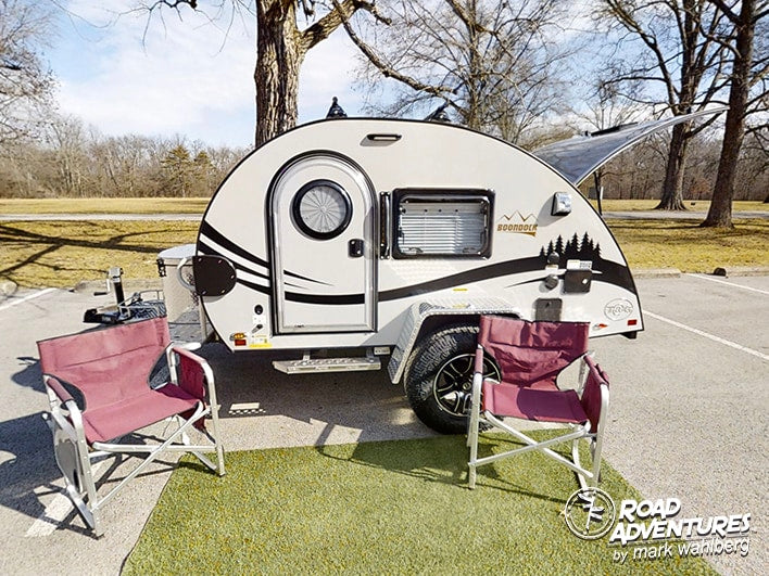 A compact teardrop camper rv rental with an open hatch, camp chairs, and mat, ready for a cozy outdoor adventure.