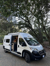 White Winnebago Solis RV rental with side door open, showing its interior, parked in a shaded wooded area.