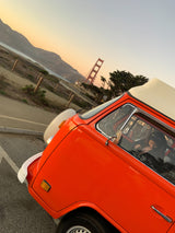 A vibrant orange VW campervan, perfect for an RV rental, is parked near the Golden Gate Bridge at sunset with a person inside.