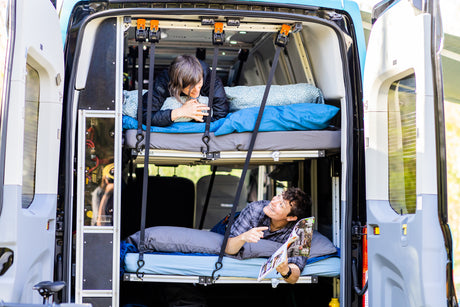 Two people relaxing on bunk beds inside an RV rental van, seen through its open rear doors. Perfect for camping.