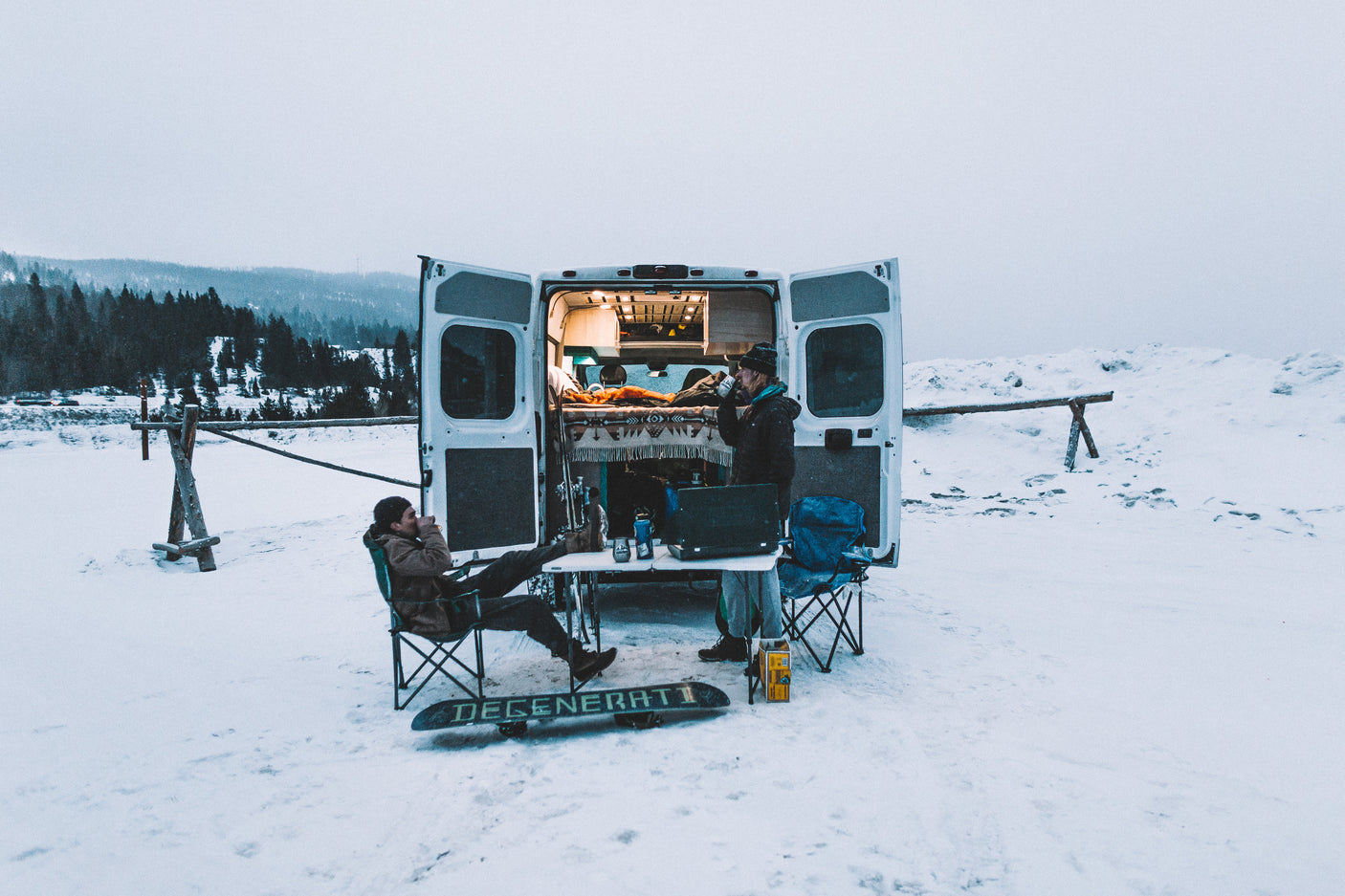 Winter RV rental adventure: Two people relax outside a camper van in snowy mountains; a snowboard lies nearby.