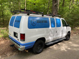 White Adventure Rig Rentals van with blue window graphics and roof rack, parked in a forest, perfect for RV rental adventures.