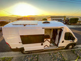 White RV rental camper van parked on a street at sunset. Its open door reveals a kitchen, small toilet, and solar panels on top.