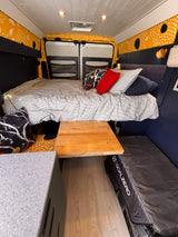 RV rental: Camper van interior with a cozy striped bed, wood table, speckled countertop, and unique yellow patterned walls.