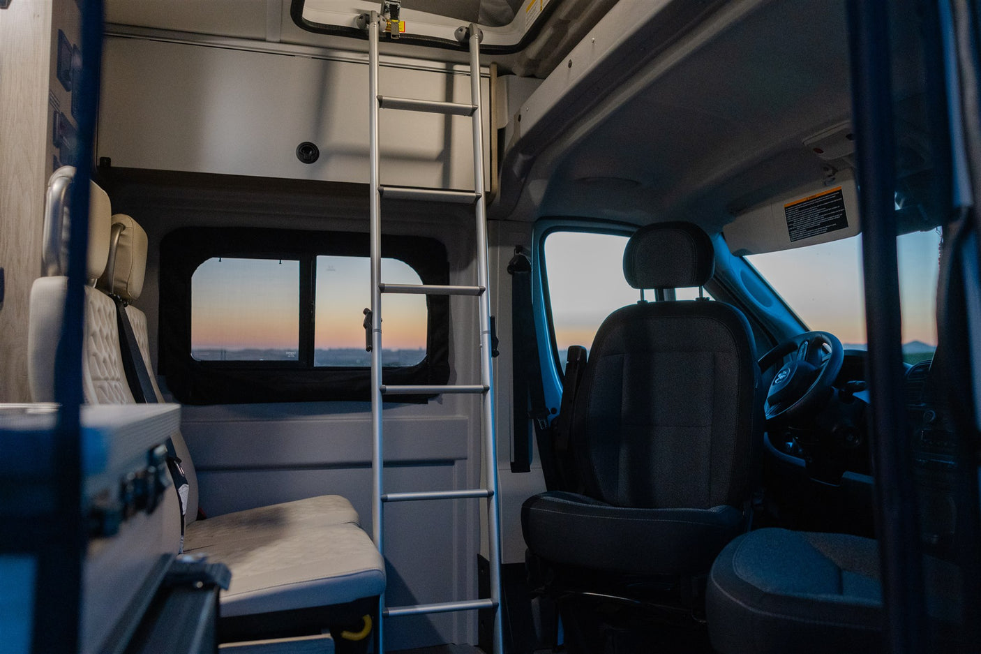 Inside an RV rental, a ladder leads to an upper bunk. Plush seating and a window with a sunset view.
