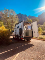 White RV rental van with open back doors, showing interior, parked on gravel amidst trees & mountains under bright sun.