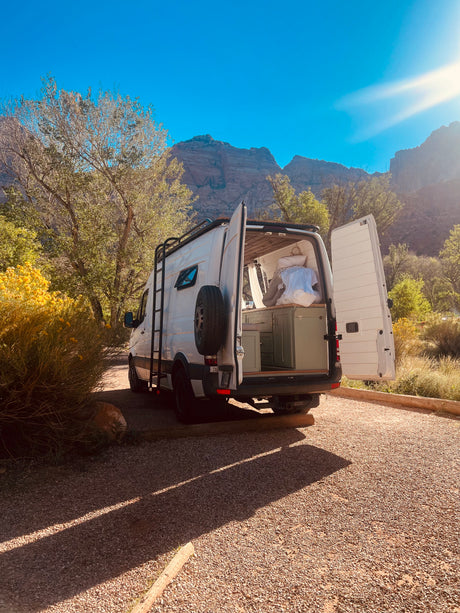 White RV rental van with open back doors, showing interior, parked on gravel amidst trees & mountains under bright sun.