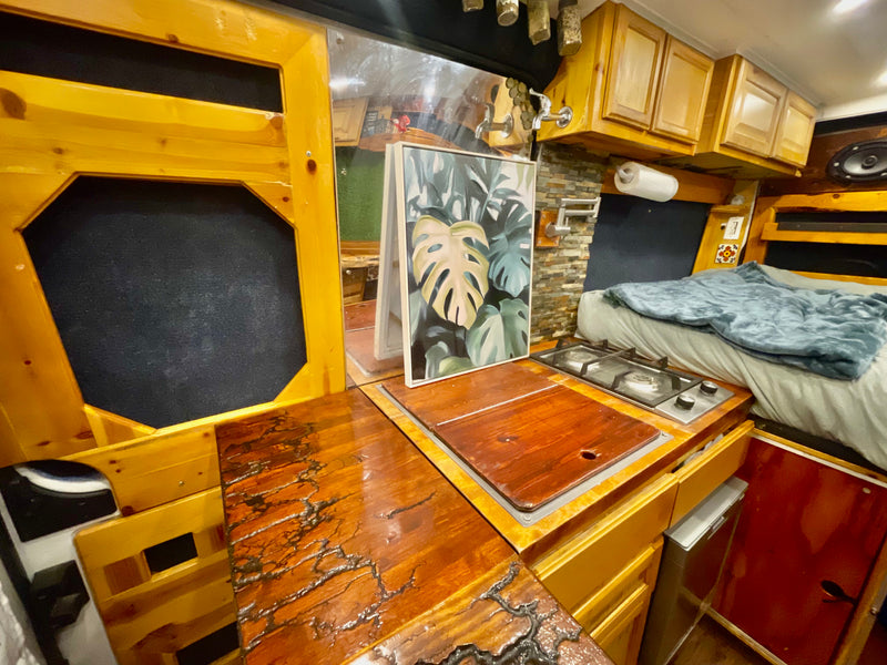 RV rental's custom wood interior: kitchen with stove & covered sink, cozy bed, plant art, unique fractal wood counter. — photo 12