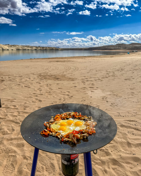 RV rental trip: Outdoor cooking setup with eggs, mushrooms, and peppers on a griddle at a sandy lake beach under blue skies.