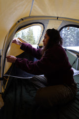 Inside a cozy tent, a person adjusts the window, enjoying the snowy camp view. Enhance your outdoor adventure with an RV rental!