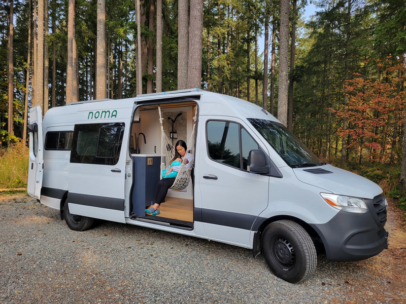 A woman with a baby rests in a hanging chair inside a white NOMA RV rental campervan, parked in a forest. — photo 7