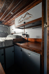 Modern RV rental interior: cozy bed, dark wood kitchen with sink, faucet, built-in fridge. Mountain-themed overhead decor.