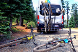 White RV rental van with bikes on a rack and two mountain bikes on the ground, set in a peaceful forest.