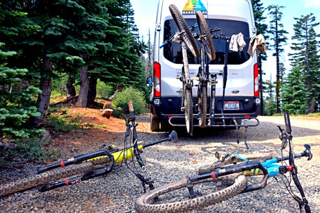 White RV rental van with bikes on a rack and two mountain bikes on the ground, set in a peaceful forest.