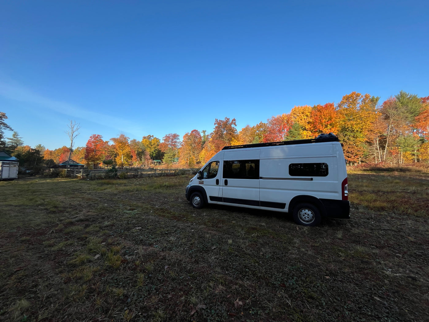 White RV rental parked on a grassy field against a vibrant autumn forest and clear blue sky, ideal for nature getaways.