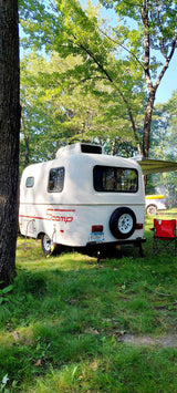 A compact Scamp RV, perfect for an RV rental, is parked in a lush, wooded campground. A red chair awaits by the back.
