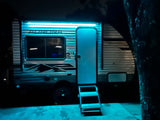 An rv rental brightly lit at night with glowing blue awning lights, an open door, and accessible entry steps on a paved surface.