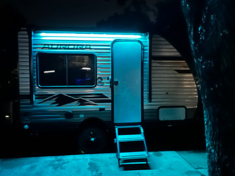 An rv rental brightly lit at night with glowing blue awning lights, an open door, and accessible entry steps on a paved surface.