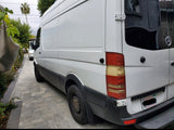Rear-side view of a white Mercedes-Benz Sprinter van, an RV rental, on a paved driveway.