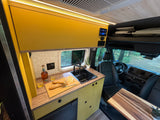 Stylish RV rental interior: yellow kitchen cabinets, sink, countertop meal setup, and driver's area with forest views.