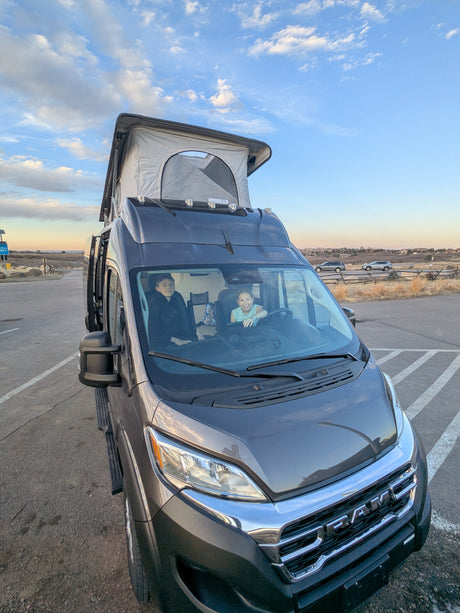 Dark grey RV rental with a pop-top roof raised, parked outdoors with two happy kids visible inside the front.