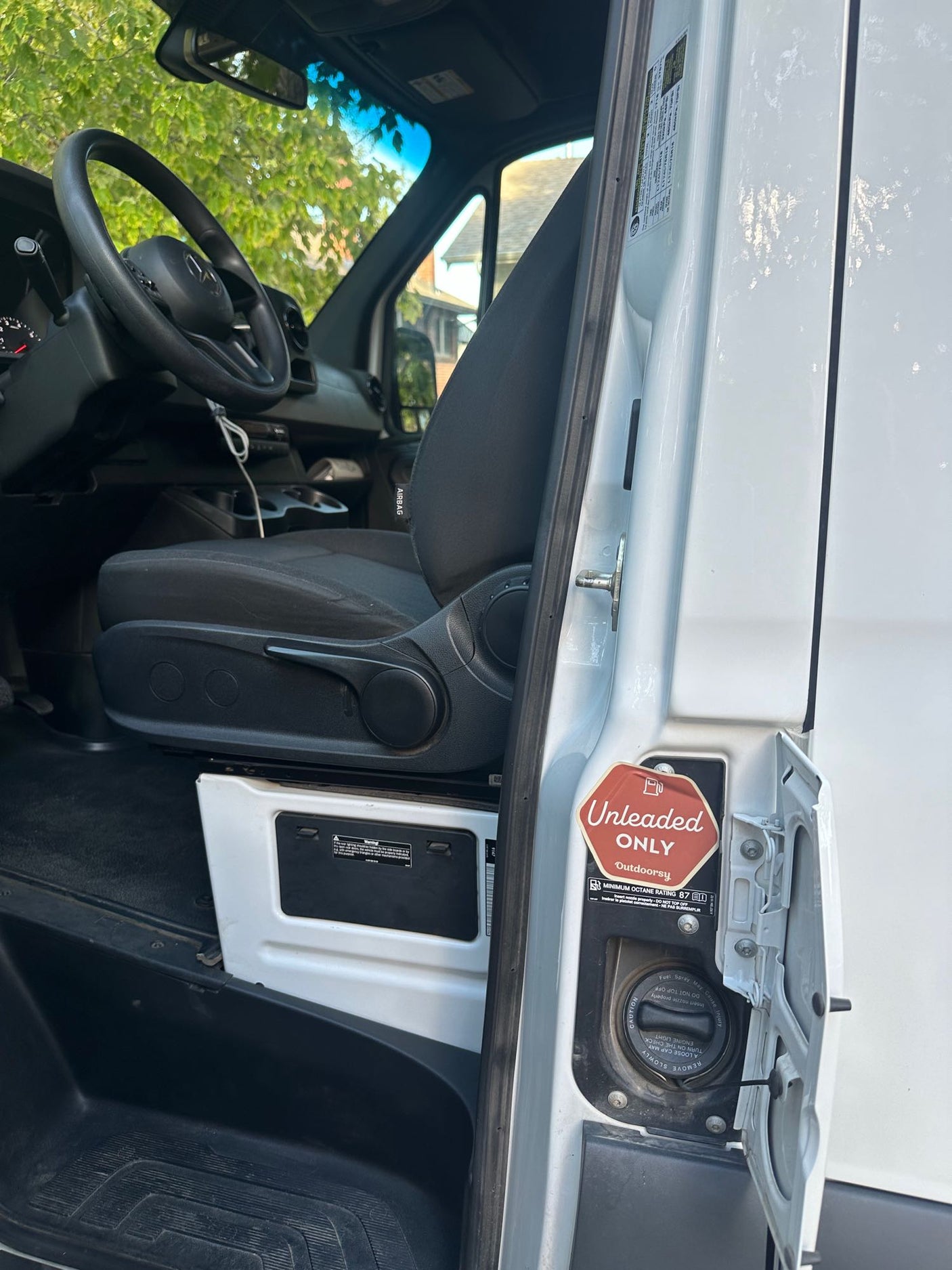 Inside a white van, the open driver's side fuel door shows "Unleaded ONLY" and fuel instructions, typical for an RV rental.