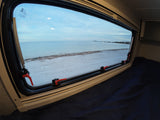 View from an RV rental: a bed with dark blue sheets, looking out a window to a serene beach, calm ocean, and distant bridge.