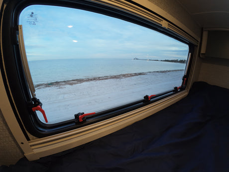 View from an RV rental: a bed with dark blue sheets, looking out a window to a serene beach, calm ocean, and distant bridge.