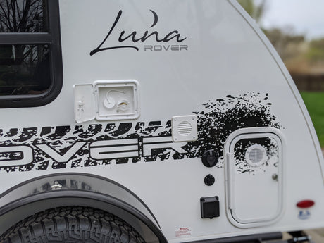 Side view of a white Luna Rover RV rental, showcasing its branding, utility hookups, and durable tire.