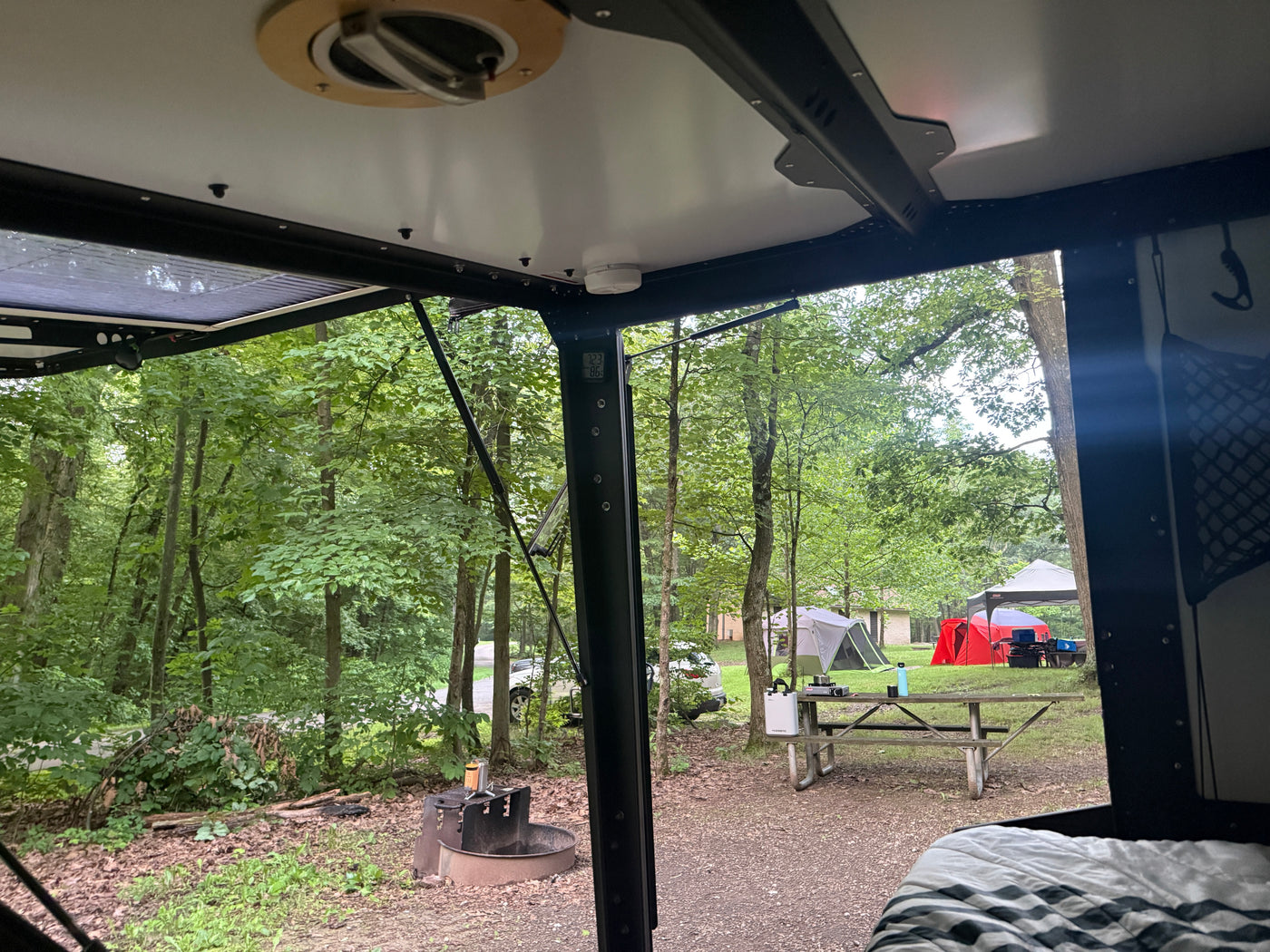 Relaxing inside an RV rental, looking out at a peaceful forest campsite with tents, a picnic table, and a fire pit. — photo 9