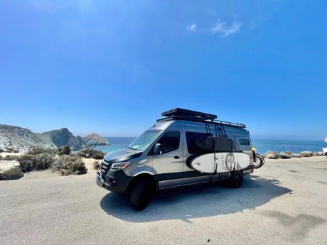 Coastal adventure awaits! Modern rv rental camper van with surfboards & bike overlooking the ocean.