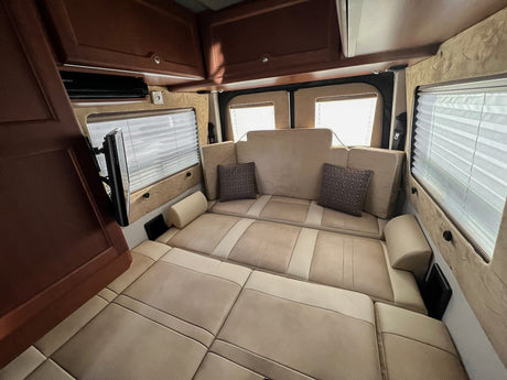 Spacious RV rental interior with a beige bed/lounge area, windows with blinds, and a mounted TV.