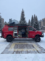 Red customized 4x4 rv rental van in snow. One person sips coffee inside, another on the roof, with a mat out.