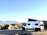 White RV rental van with pop-top roof atop a desert hill, overlooking rugged mountains under a clear sky.